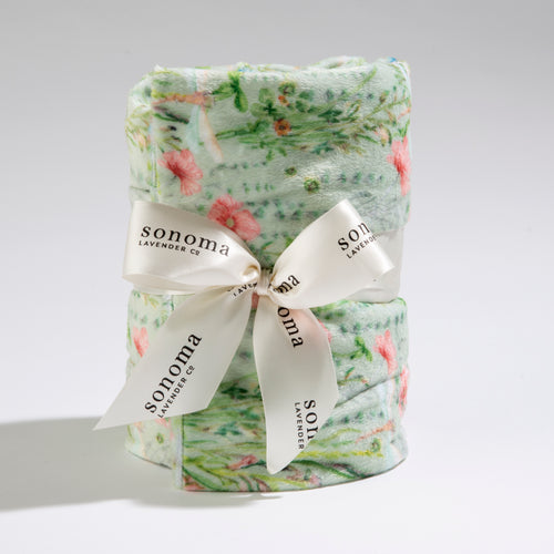 A rolled-up, soft fabric featuring a floral pattern with green and pink colors. The Sonoma Eucalyptus Dragonfly Garden Heat Wrap is neatly tied with a white ribbon that has "sonoma lavender co." printed on it in black text. The background is plain white. This heat wrap is part of the Hampton Court Essential Luxuries & The Lavender Shop collection.