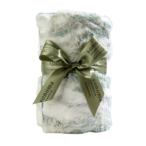 The Sonoma Lavender Sonoma Eucalyptus Snowmint Heat Wrap is a fluffy, white and light green faux fur blanket rolled up and tied with a "Sonoma Eucalyptus" green ribbon, offering cozy aromatherapy comfort.