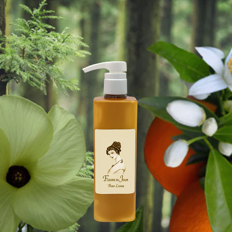 A bottle of La Bouquetiere Fleurs du Jour / Marina Blue Body Lotion with a pump stands before green leaves, white flowers, an orange, and tree trunks. The label features a woman's profile and notes its aloe moisturizer and lasting fragrance.