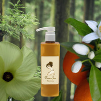 A bottle of La Bouquetiere Fleurs du Jour / Marina Blue Body Lotion with a pump stands before green leaves, white flowers, an orange, and tree trunks. The label features a woman's profile and notes its aloe moisturizer and lasting fragrance.