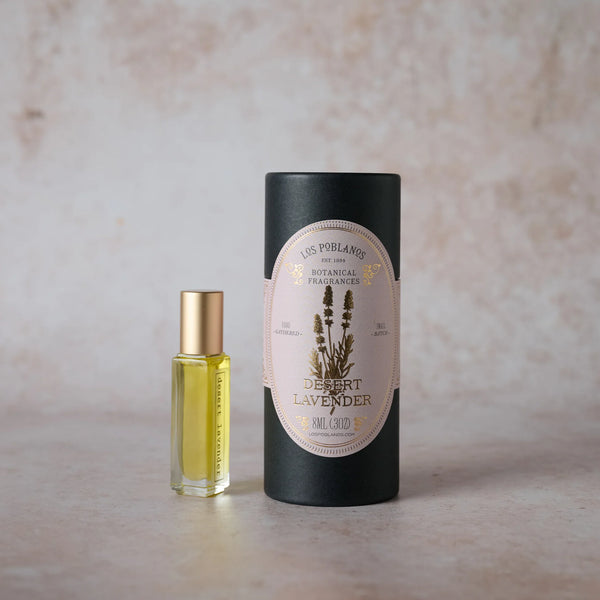 A small glass bottle of Los Poblanos Lavender Desert Botanical Fragrance with a gold cap sits beside a tall black Los Poblanos container, both featuring organic jojoba seed oil, on a light textured surface.