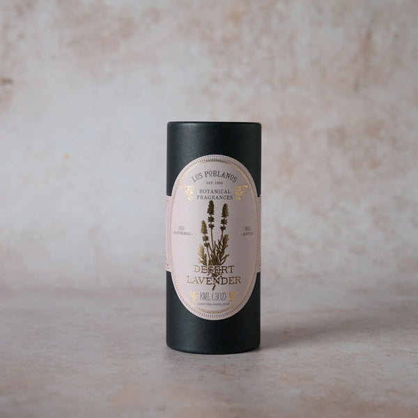 A cylindrical black container of Los Poblanos Lavender Desert Lavender Botanical Fragrance stands upright on a light, textured surface. The pastel label features lavender illustrations, gold accents, and is infused with lavender essential oil.