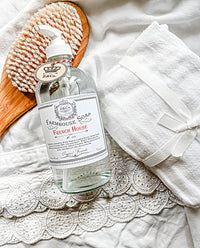 A pump bottle of Z&Co. French House Farmhouse Liquid Soap with a lavender scent sits on a lace-trimmed white cloth, accompanied by a wooden brush with bristles and a white towel tied with a ribbon. The scene gives an elegant, vintage feel with a focus on cleanliness and self-care.