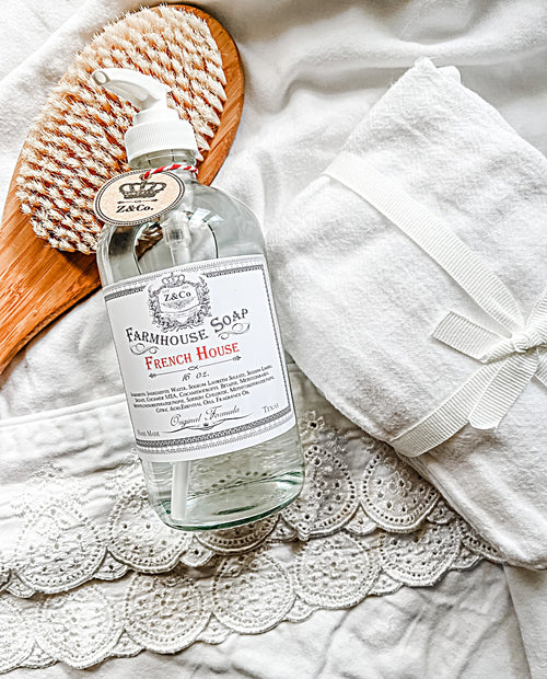 A pump bottle of Z&Co. French House Farmhouse Liquid Soap with a lavender scent sits on a lace-trimmed white cloth, accompanied by a wooden brush with bristles and a white towel tied with a ribbon. The scene gives an elegant, vintage feel with a focus on cleanliness and self-care.