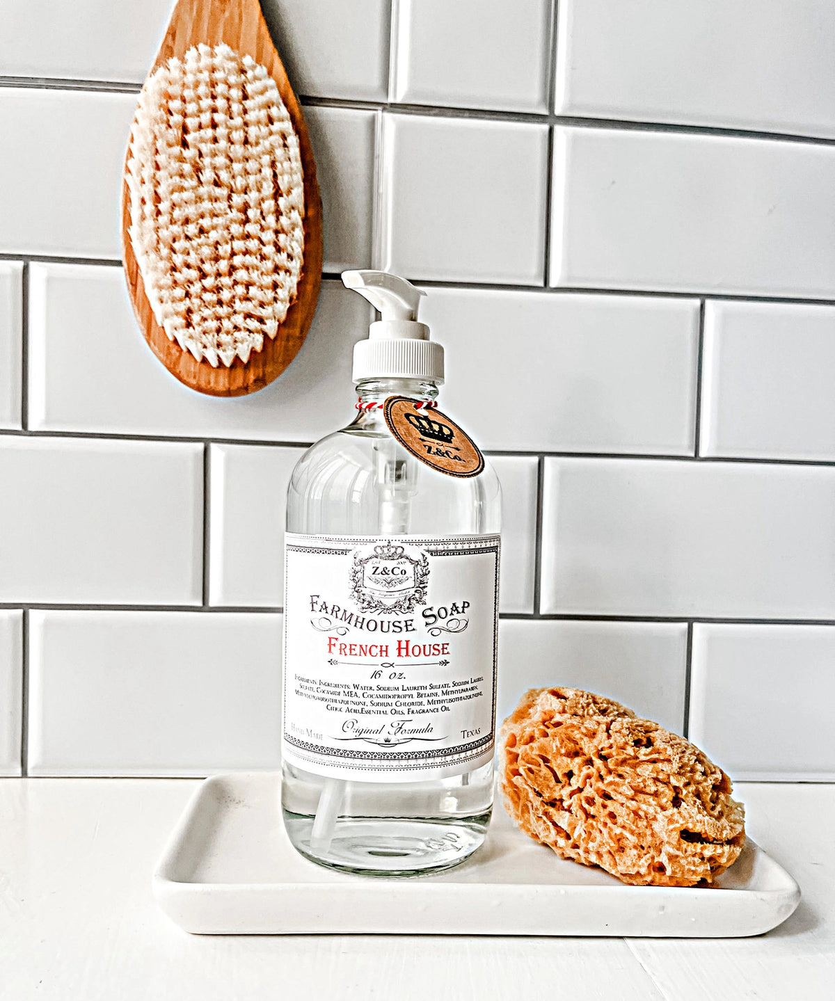 A bottle of Z&Co. French House Farmhouse Liquid Soap with a lavender scent sits on a white tray in front of a tiled wall. Next to it is a natural sea sponge, and above, a wooden bath brush hangs on the wall. The scene exudes a clean, simple, and elegant bathroom aesthetic reminiscent of a French House.