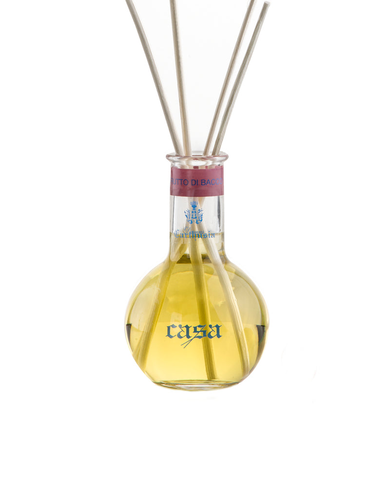 A transparent Carthusia Frutto di Bacco Reed Diffuser with a bulbous base containing yellow liquid, labeled "rosa." It has several reeds inserted into the neck, against a plain white background and emanates fragrant.