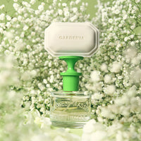 A 60ml Caswell Massey Gardenia Eau de Toilette bottle with a green and white cap is surrounded by delicate baby's breath, highlighting this floral fragrance against a light green background.