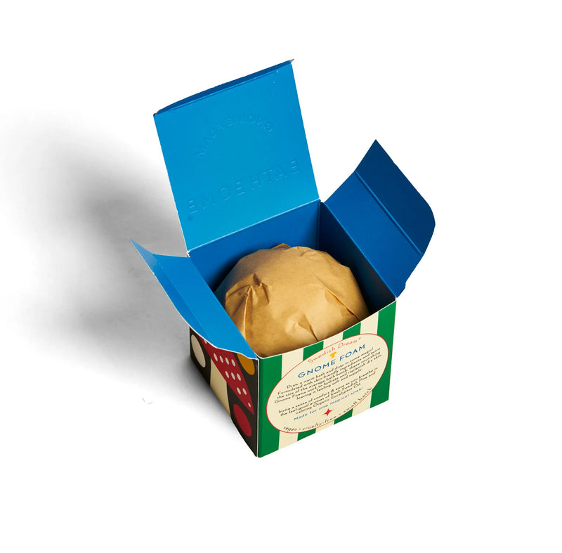 A blue, green, and white box with colorful dots and a circular label holds the Kala Swedish Dream Gnome Foam™ Swedish Pine Bath Bomb, wrapped in brown paper inside under the open lid.