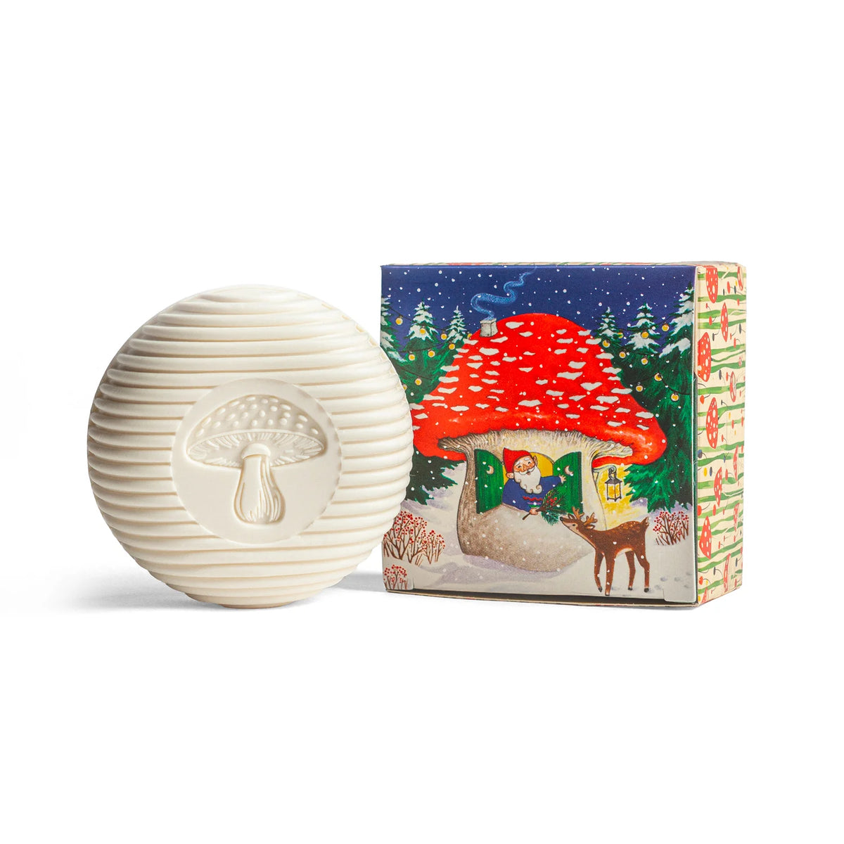 A circular bar of Kala's Swedish Dream Gnome Soap, showcasing a mushroom motif, rests alongside a vibrant package that depicts a gnome house, an elf, and a deer within an enchanting snowy forest filled with Swedish pine.