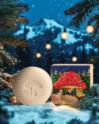 A round, white bar of Swedish Dream Gnome Soap by Kala, featuring a mushroom engraving, rests on snow next to a colorful box showcasing a red-roofed house. The scene is framed by evergreen branches and warm lights, enhanced by the Swedish Pine fragrance, with snow-covered mountains in the background.