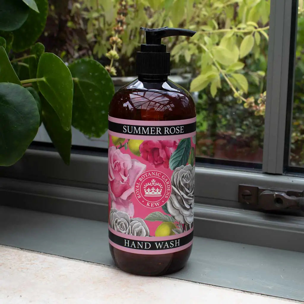 A bottle of The English Soap Co. Kew Gardens Summer Rose Hand Wash with a floral label sits on an indoor windowsill beside a green leafy plant, with more greenery visible outside the window.
