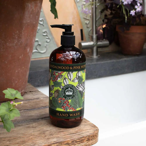 A bottle of The English Soap Co. Kew Gardens Sandalwood and Pink Pepper Hand Wash rests on a wooden surface beside a bathtub, with potted plants and flowers in the background.