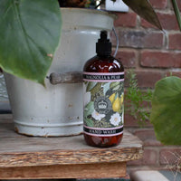 A bottle of The English Soap Co. Kew Gardens Magnolia and Pear Eau de Toilette rests on a wooden surface next to a white metal pot and green leaves, with a brick wall in the background.