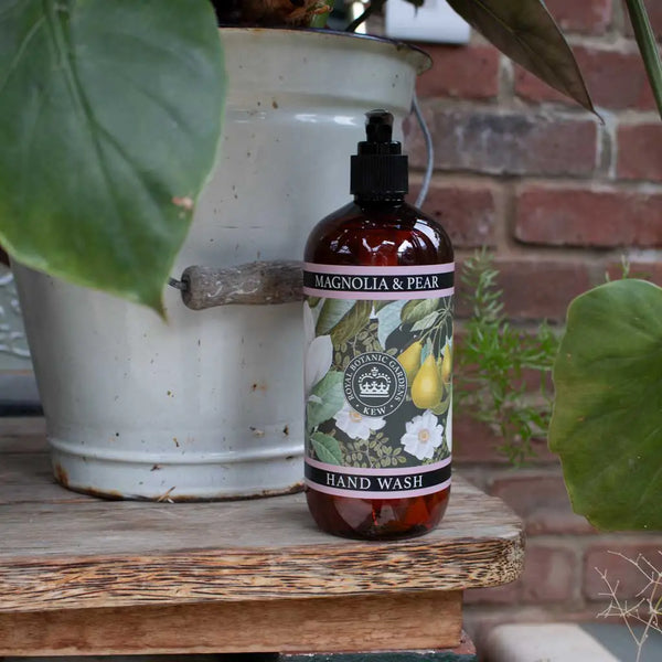 A bottle of The English Soap Co. Kew Gardens Magnolia and Pear Hand Wash sits on a wooden surface beside a white metal pot with large green leaves, set against a brick wall background.