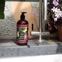 A bottle of The English Soap Co. Kew Gardens Osmanthus Rose Hand Wash rests on a gray counter beside a silver faucet, with decorative tiles and purple flowers in a brown pot surrounding it.