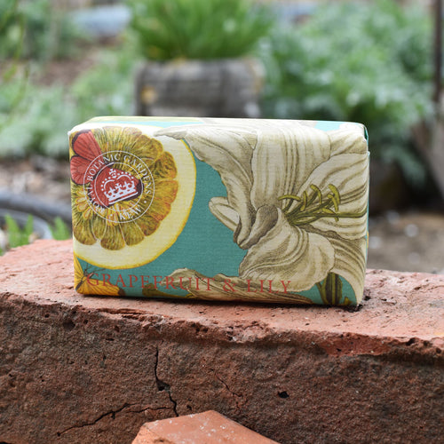 The English Soap Co. Kew Gardens Grapefruit and Lily Soap, wrapped in decorative paper with grapefruit and lily illustrations, rests on a red brick outdoors among greenery.
