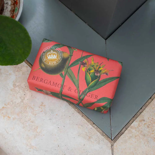 A red rectangular bar of The English Soap Co. Kew Gardens Bergamot and Ginger Soap, adorned with floral illustrations and the text "Bergamot & Ginger" on the wrapper, rests on a tiled surface. The packaging also showcases a circular label with a crown emblem and the text "BOTANIC GARDENS KEW." A green leaf is partially visible, hinting at a luxurious bathing experience.