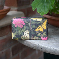 The English Soap Co. Kew Gardens Osmanthus Rose Soap, a vegan-friendly bar wrapped in floral paper with butterflies and leaves, rests on stone near potted plants and a brick wall.