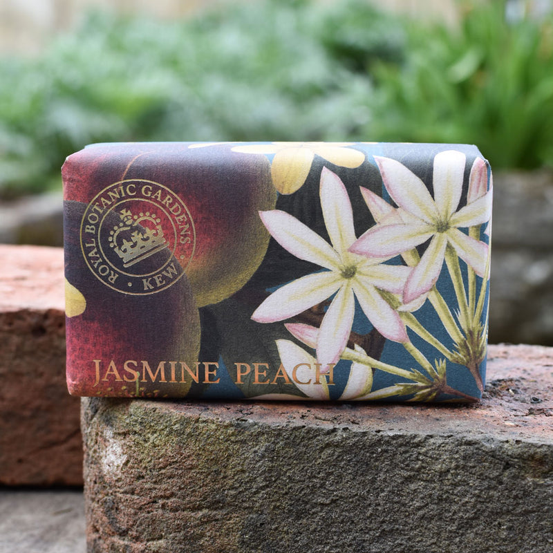 A bar of The English Soap Co. Kew Gardens Jasmine Peach Soap, wrapped in floral paper with the brand's seal, rests on a brick surface outdoors amid lush greenery.