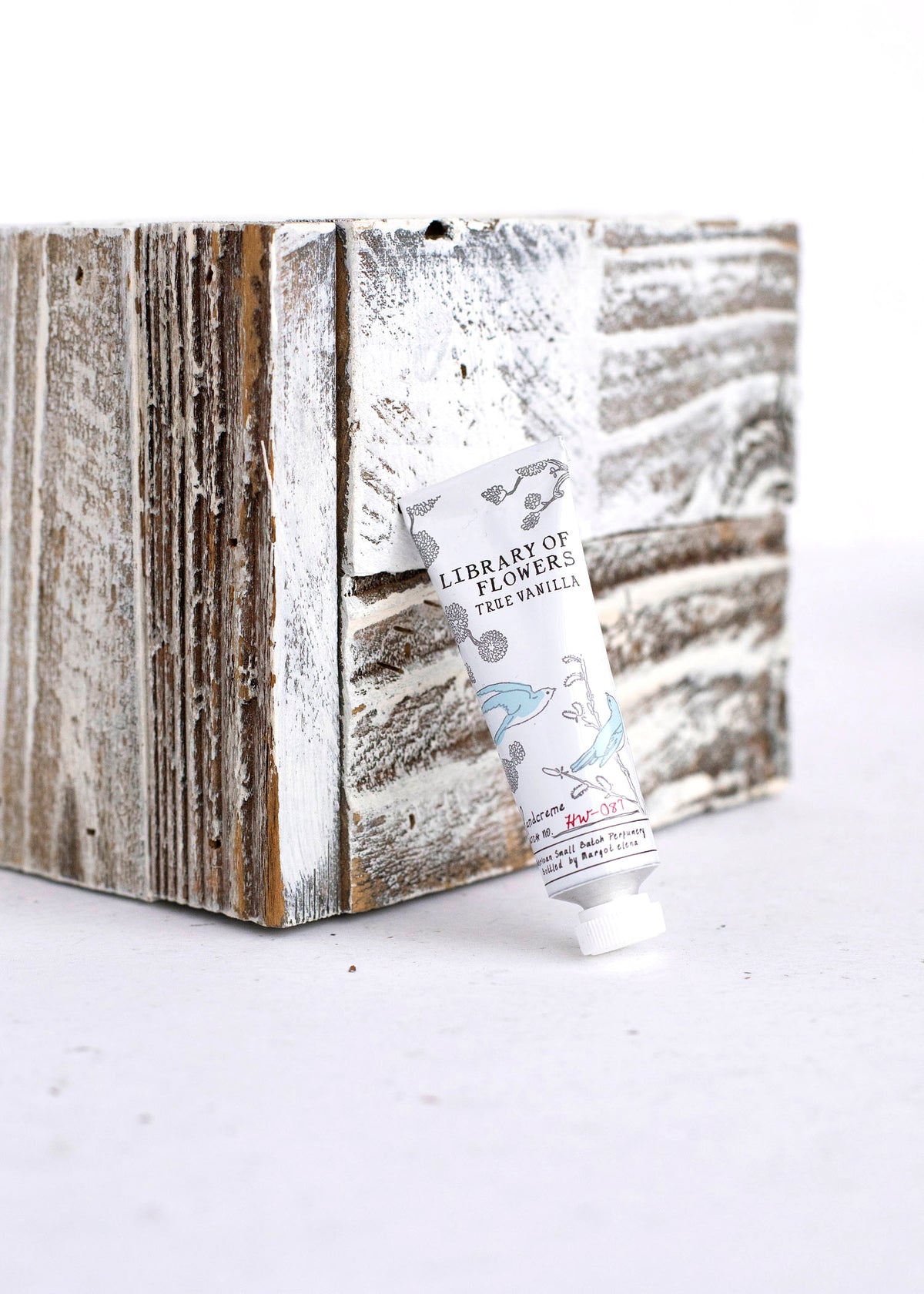 A Library of Flowers True Vanilla Petite Treat Handcreme hand cream tube by Margot Elena with deep hydration leans against a rustic wooden block with a distressed white paint finish, against a light background.