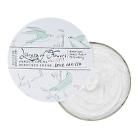 A round container of Margot Elena brand Library of Flowers True Vanilla Parfum Crema. The lid features elegant illustrations of hummingbirds and plants, with a white cream visible inside the open container.