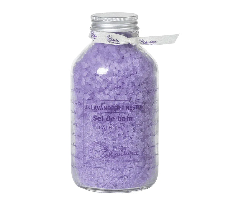 A clear glass bottle filled with Lothantique les lavandes de l'oncle Nestor lavender bath salts, labeled "Lothantique, sel de bain, bath salt, enchantique Lothantique" with a white ribbon tied around.