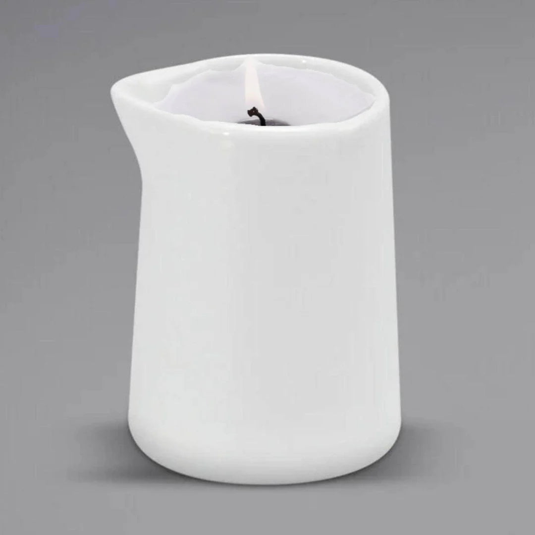 A lit Sonoma Lavender Massage Candle by Sonoma Lavender is placed in a white ceramic pitcher candle holder with a small spout, set against a plain gray background for spa-quality relaxation.