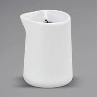 A lit Sonoma Lavender Massage Candle by Sonoma Lavender is placed in a white ceramic pitcher candle holder with a small spout, set against a plain gray background for spa-quality relaxation.