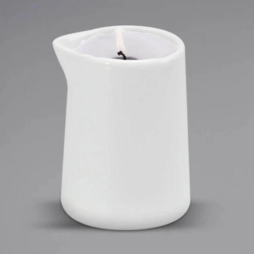A lit Sonoma Lavender Massage Candle by Sonoma Lavender is placed in a white ceramic pitcher candle holder with a small spout, set against a plain gray background for spa-quality relaxation.