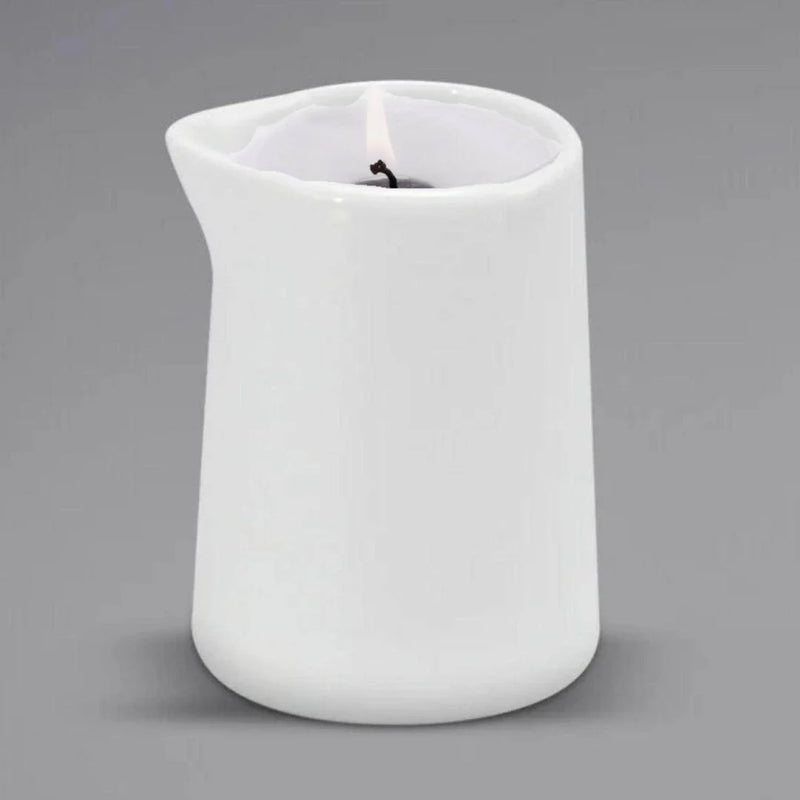 A lit Sonoma Lavender Massage Candle by Sonoma Lavender is placed in a white ceramic pitcher candle holder with a small spout, set against a plain gray background for spa-quality relaxation.