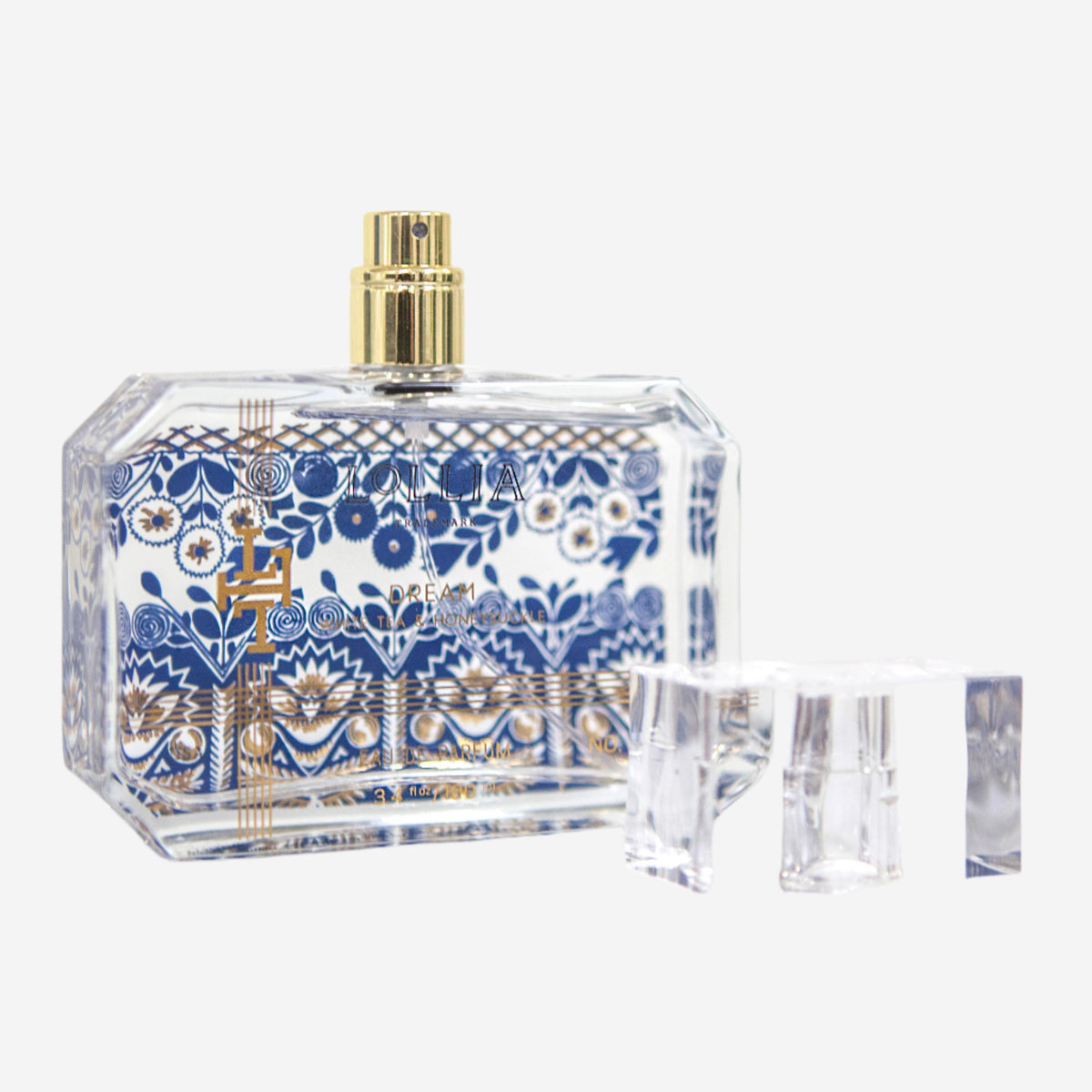 A clear glass Margot Elena Lollia Dream Eau de Parfum bottle with a gold spray top and blue and gold floral accents, featuring Honeysuckle and White Tea notes, sits next to its transparent cap on a white background.