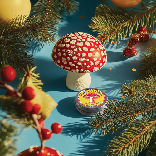 A realistic red and white spotted mushroom model stands on a blue surface with pine branches, red berries, yellow ornaments, and a tin of Kala Swedish Dream Minty Mushroom Lip Balm, made with organic mushroom extracts for natural lip care.