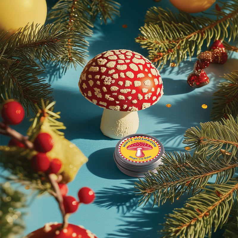 A realistic red and white spotted mushroom model stands on a blue surface with pine branches, red berries, yellow ornaments, and a tin of Kala Swedish Dream Minty Mushroom Lip Balm, made with organic mushroom extracts for natural lip care.