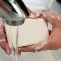 Hands washing with a bar of The English Soap Co. Sandalwood Vintage Italian Wrapped Soap under running water from a faucet, with soap bubbles visible on the hands and soap.
