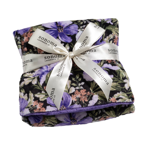 A folded blanket with a purple floral design is tied with a white ribbon decorated with "Sonoma Lavender" text. The fabric features a pattern of lavender and green leaves, giving it a lush, botanical appearance, reminiscent of the calming Sonoma Lavender Midnight Garden Heat Wrap.