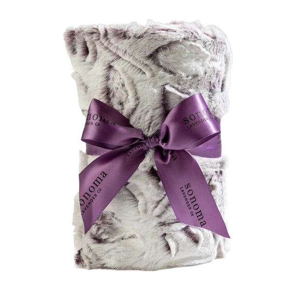 A rolled-up faux fur blanket with a white and gray pattern, tied with a purple "Sonoma Lavender" ribbon, offers the same cozy luxury as the Sonoma Lavender Iced Plum Heat Wrap for ultimate relaxation and comfort.