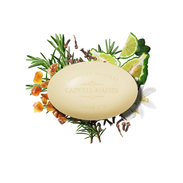 The Caswell-Massey Number Six Bar Soap, a citrus woody-scented oval soap by Caswell-Massey, is shown with rosemary, lime slices, cloves, amber resin, and white flowers on a white background.