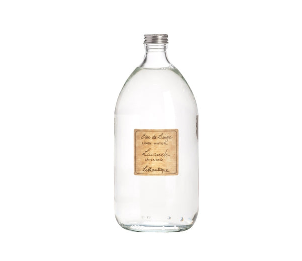 A clear glass bottle with a silver cap, filled with a transparent liquid. It has a beige label affixed to the front, featuring text in a stylish font that reads "Lothantique Lavender Linen Water Spray - 1 liter/33oz." This scented water is perfect for use with your steam iron to refresh linens. The bottle is placed against a white background.