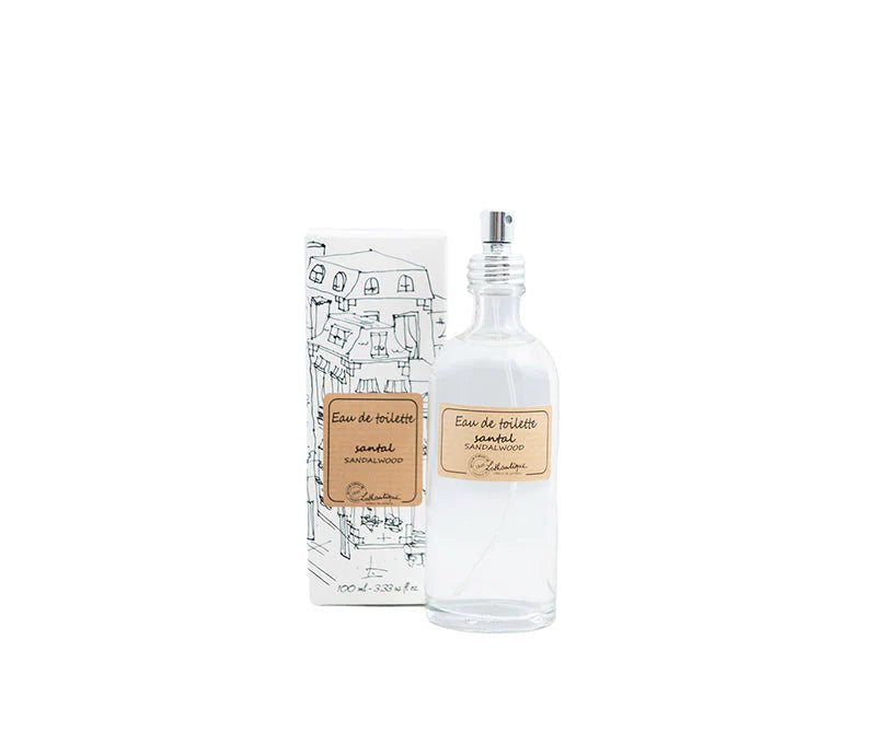 A clear glass perfume bottle with a pump, labeled "Lothantique Sandalwood Eau de Toilette," is positioned next to a box decorated with a line drawing of houses. This personal fragrance bottle embodies simplicity and elegance, showcasing a neutral design that suits any decor.