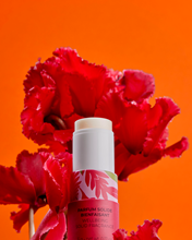 The Roger & Gallet Red Ginger Wellbeing Solid Perfume, in a red and pink-labeled tube, stands before bold red flowers and an orange background—an alcohol-free fragrance with natural ginger extract from Roger & Gallet.