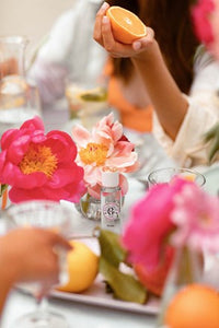 A person holding a halved Roger & Gallet Rose orange at a beautifully decorated table with bright pink and damask rose flowers and several oranges.