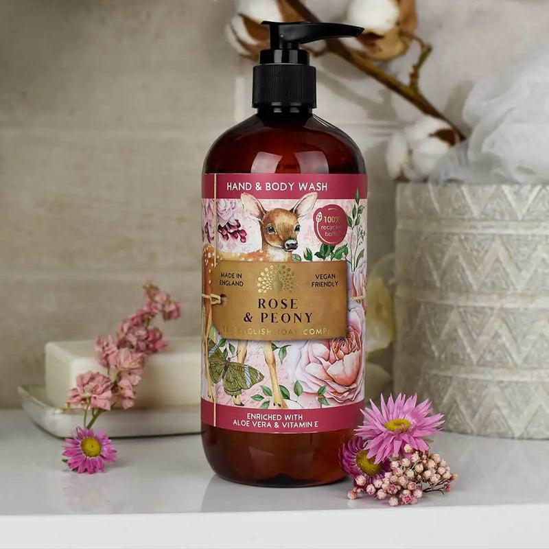 A bottle of "The English Soap Co. Anniversary Rose & Peony Hand & Body Wash" is displayed next to pink flowers. The bottle features a colorful design with images of roses, peonies, and a deer. The product is described as vegan-friendly, made in England, and enriched with aloe vera and vitamin E.