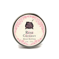 A tin labeled "Three Sisters Aphothecary Rose Coconut Body Butter" (4 oz/113g) with a pink floral border highlights this natural skincare shea butter moisturizer against a white background.