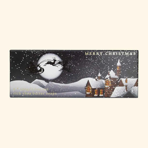 The Winter Village Triple Soap Gift Box by The English Soap Co. displays a charming snowy scene featuring illuminated houses and a large moon. Across the starry sky, Santa and his reindeer make their journey. This rectangular box is adorned with the phrases "Merry Christmas" and "rich shea butter scented soaps," making it an ideal Christmas present.