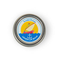 A round tin container labeled "Swedish Dream Lemon Lip Balm" promises clean beauty and long-lasting hydration. The label features an illustration of a red sailboat on the water against a yellow background, with the text "net wt. 0.8 oz / 22.9g" at the bottom.