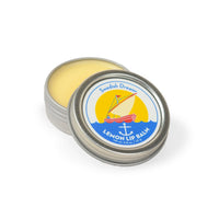 A small, opened tin of Swedish Dream Lemon Lip Balm with a yellow balm inside. The tin lid, featuring the product label with a boat on the sea against a yellow sun and blue sky, rests beside the container. This Clean Beauty product provides long-lasting hydration and weighs 0.8 ounces (22g).