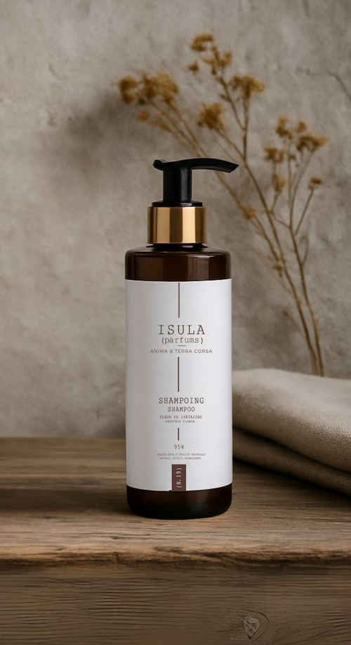 A brown and white pump bottle of ISULA parfums Fiore di Castagna Chestnut Flower Shampoo stands before beige folded towels and dried flowers, reflecting European heritage and offering a gentle cleanse against a neutral wall.