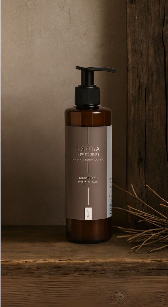 A brown pump bottle of ISULA parfums Storia di Mare - Sea Story Shampoo rests on a wooden surface with a rustic backdrop and dried twigs. The label displays "ISULA (PAE FIUMA) ANIMA E TERRA CORSA SHAMPOING RIVOLI DI MARE.