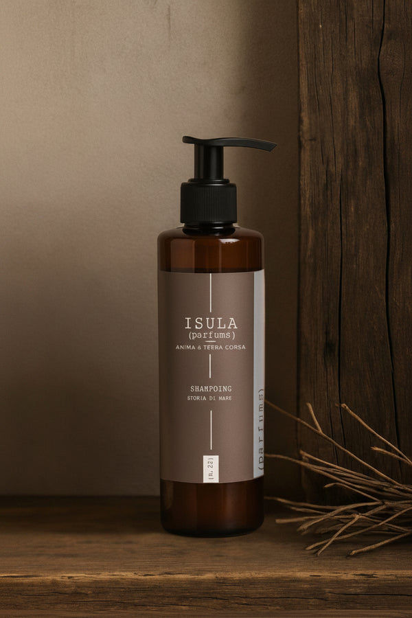 A brown pump bottle of ISULA parfums Storia di Mare - Sea Story Shampoo rests on a wooden surface with a rustic backdrop and dried twigs. The label displays "ISULA (PAE FIUMA) ANIMA E TERRA CORSA SHAMPOING RIVOLI DI MARE.