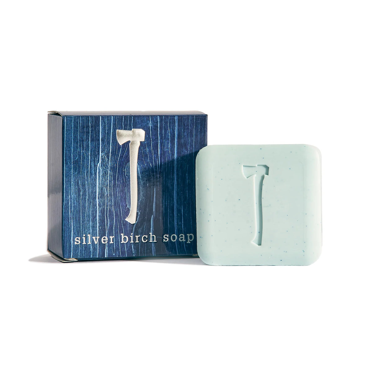 A bar of Kala's Silver Birch Bar Soap, adorned with an axe imprint, is showcased beside its packaging. The blue box displays a white axe design along with the lowercase words "silver birch soap." This light blue soap, infused with vetiver and gentle exfoliating speckles, offers a refreshing clean.